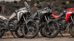 Três motocicletas Honda Sahara 300 2027 enfileiradas ao ar livre, em tons de cinza claro, cinza-escuro e vermelho