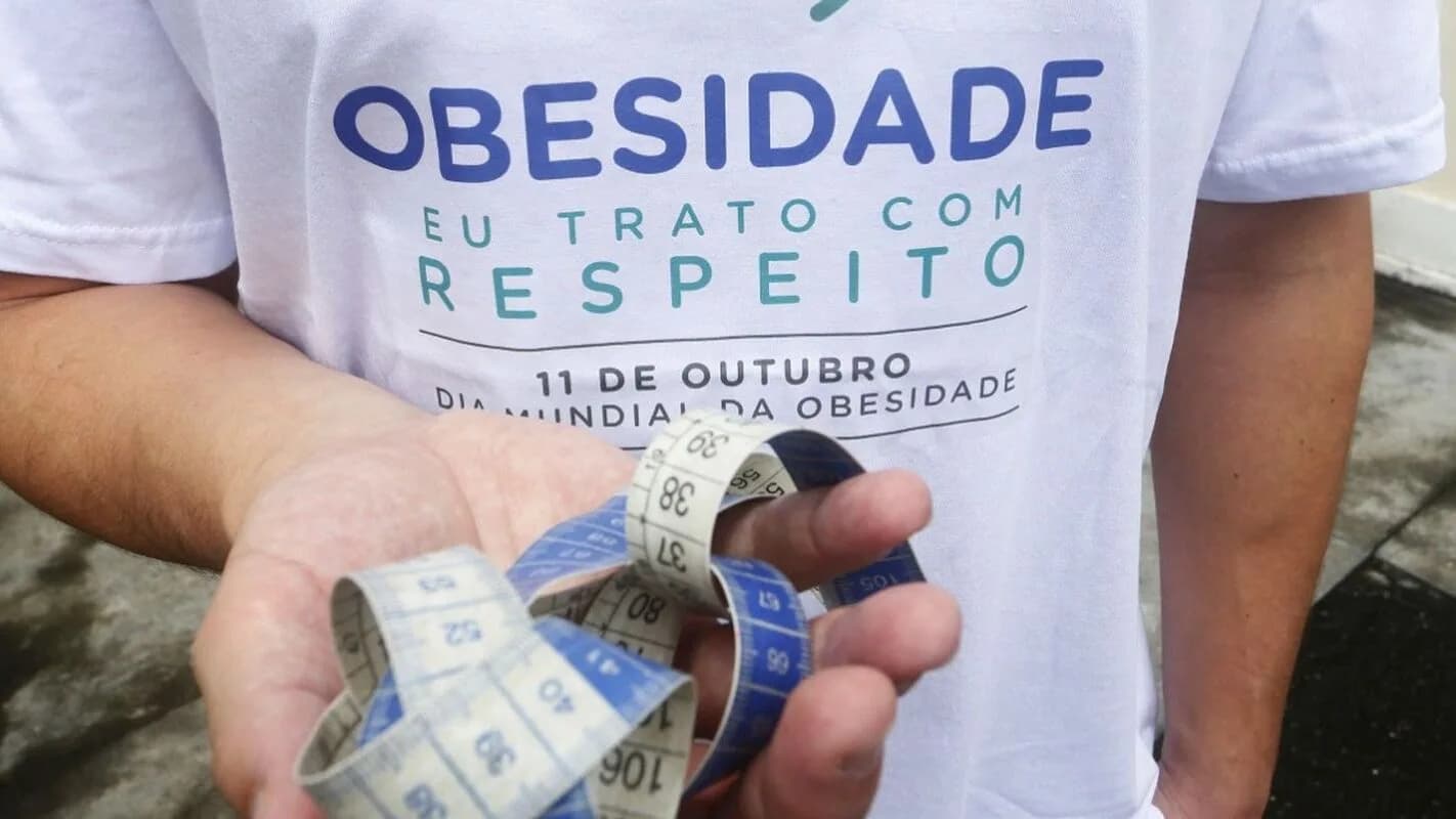 Pessoa segura fita métrica junto à camiseta com a frase 'Obesidade eu trato com respeito'