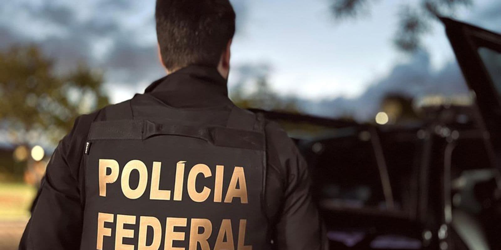 Agente da Polícia Federal de costas, com colete escrito Polícia Federal, ao lado de viatura