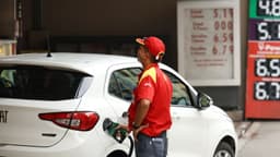 Frentista de uniforme vermelho e boné abastece um carro branco em posto de combustível, com o painel de preços ao fundo. Imagem de arquivo