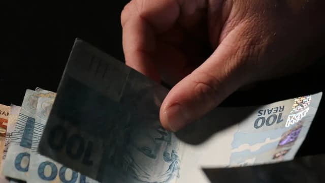 Mãos contam notas de real, ilustrando a dívida pública federal