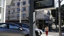 Termômetro de rua marca 42 °C em uma calçada do Rio de Janeiro, com carros estacionados e uma pedestre caminhando de costas ao fundo, em foto de arquivo