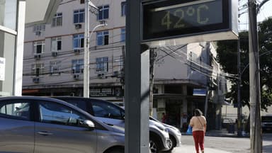 Termômetro de rua marca 42 °C em uma calçada do Rio de Janeiro, com carros estacionados e uma pedestre caminhando de costas ao fundo, em foto de arquivo
