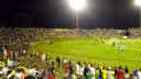 Torcida em arquibancada lotada acompanha jogo de futebol à noite sob holofotes