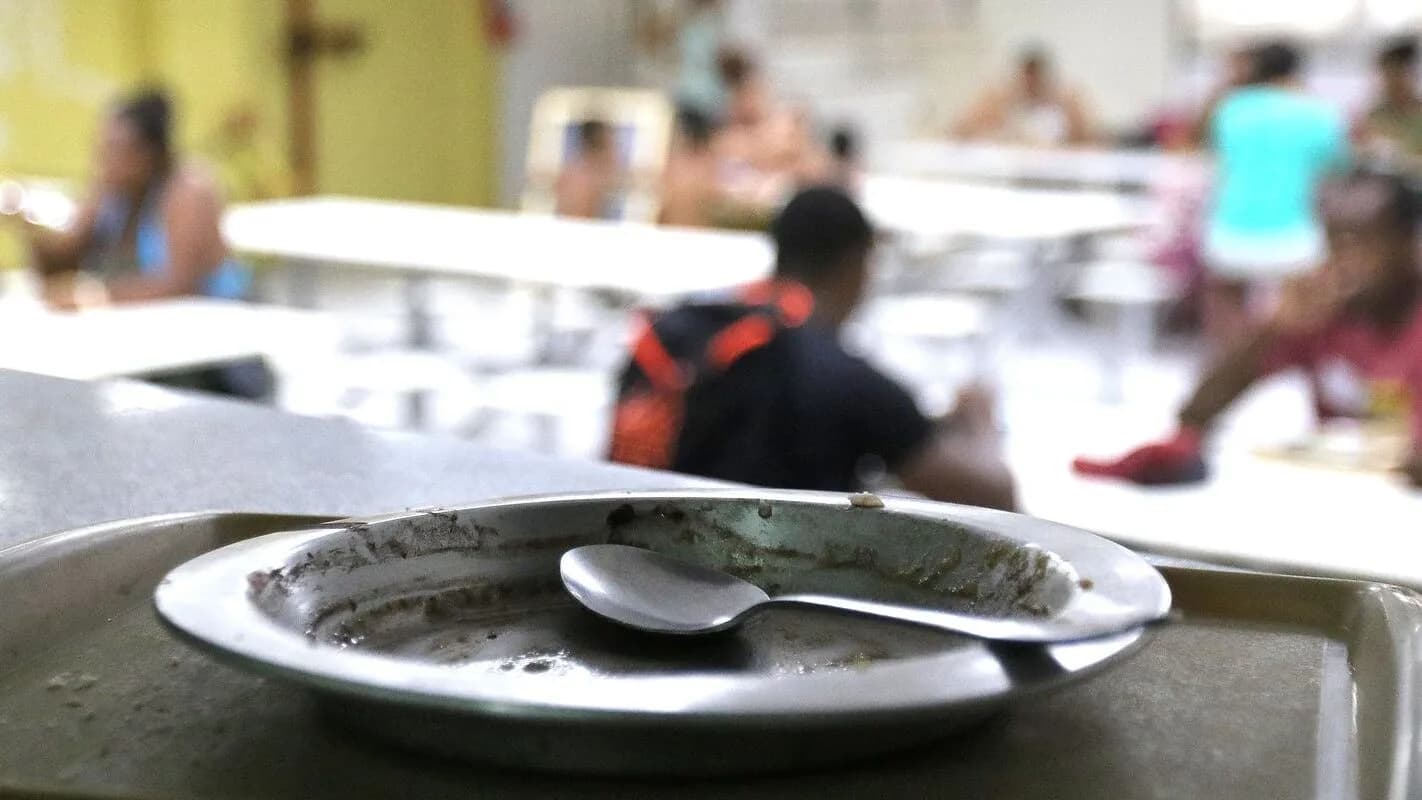 Pessoas em refeitório popular, com prato vazio em primeiro plano, ilustrando o tema da insegurança alimentar no Brasil