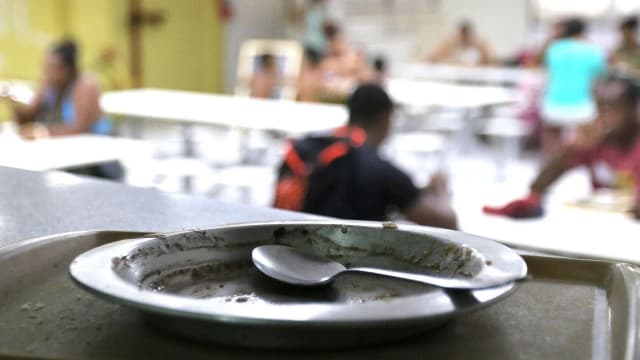 Pessoas em refeitório popular, com prato vazio em primeiro plano, ilustrando o tema da insegurança alimentar no Brasil