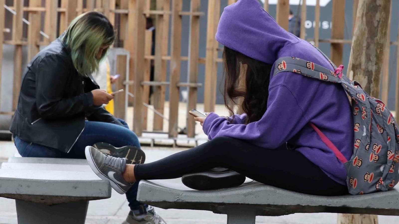 Duas jovens sentadas em bancos na rua, cada uma olhando o próprio celular com a cabeça inclinada para baixo; imagem de arquivo