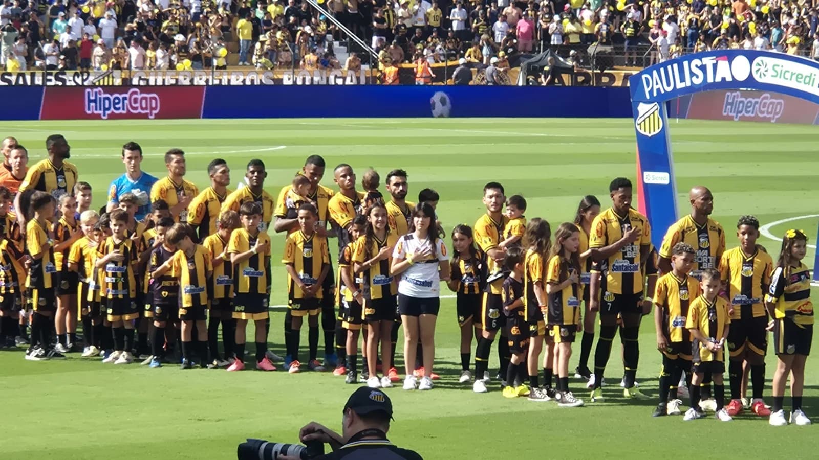 Jogadores do Grêmio Novorizontino perfilados no gramado, uniforme amarelo e preto, antes de partida no Estádio Jorge Ismael de Biasi
