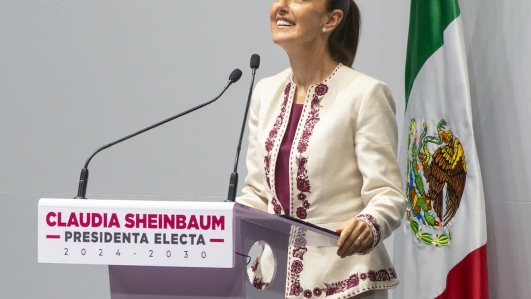 Claudia Sheinbaum discursa em evento oficial, com a bandeira do México ao fundo