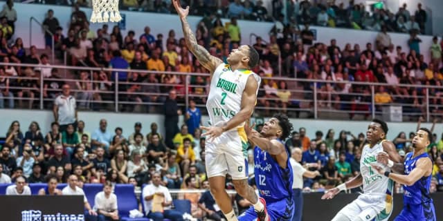 Jogador da seleção brasileira de basquete arremessa ao gol durante partida das Eliminatórias, com defensor da Venezuela tentando bloquear