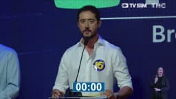 Gabriel Azevedo, candidato do MDB ao governo de Minas Gerais, de camisa branca com adesivo do número 15, fala ao microfone no púlpito do debate da TV Sim Brasil e da TMC com o cronômetro zerado na tela e intérprete de Libras ao fundo