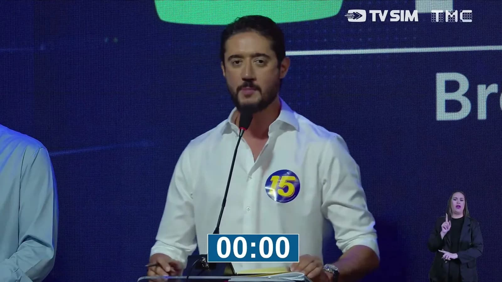 Gabriel Azevedo, candidato do MDB ao governo de Minas Gerais, de camisa branca com adesivo do número 15, fala ao microfone no púlpito do debate da TV Sim Brasil e da TMC com o cronômetro zerado na tela e intérprete de Libras ao fundo