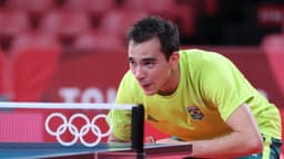Hugo Calderano, de amarelo e verde, em ação em quadra de tênis de mesa durante os Jogos Olímpicos de Tóquio 2020, com os aros olímpicos visíveis na mesa