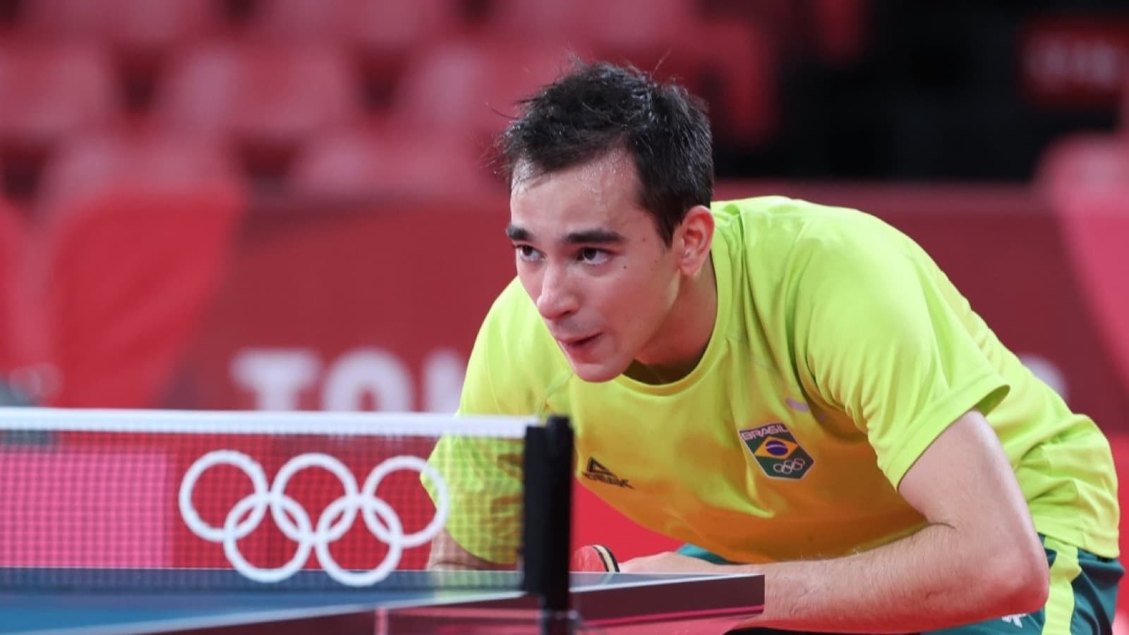 Hugo Calderano, de amarelo e verde, em ação em quadra de tênis de mesa durante os Jogos Olímpicos de Tóquio 2020, com os aros olímpicos visíveis na mesa