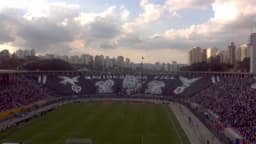 Torcida lotando arquibancada de estádio com mosaico em preto e branco durante jogo de futebol