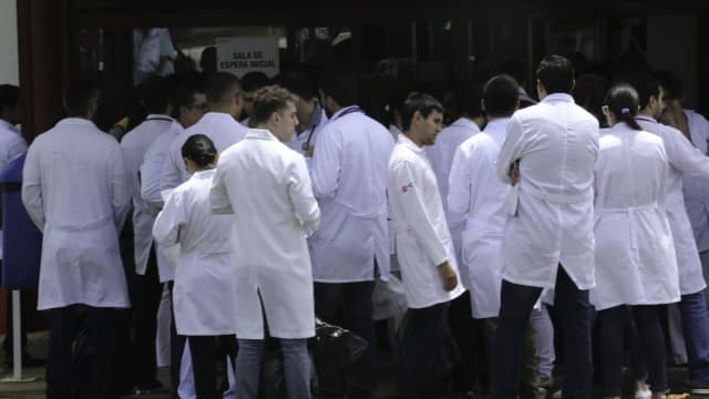 Candidatos de jaleco branco aguardam em fila para entrar em sala de prova de concurso público da área da saúde