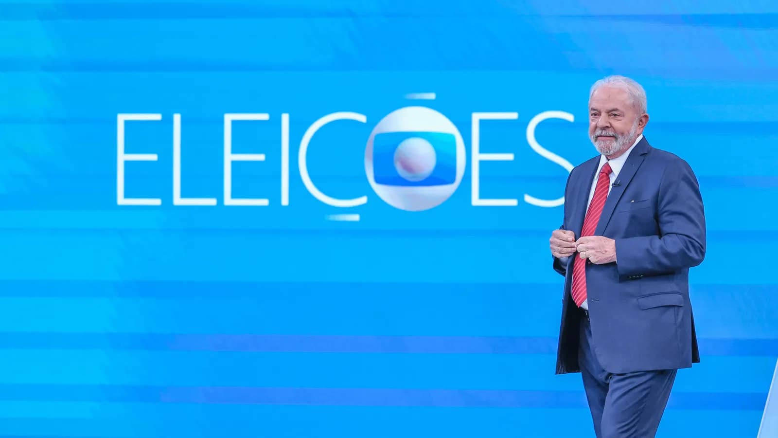 Lula em pé diante do logotipo Eleições da TV Globo em estúdio de debate presidencial anterior