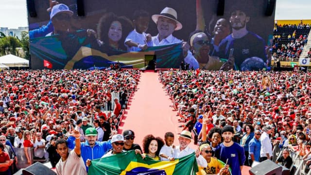 Apoiadores erguem bandeira do Brasil em frente a palanque com banner Presidente Lula Vice Alckmin no lançamento da campanha
