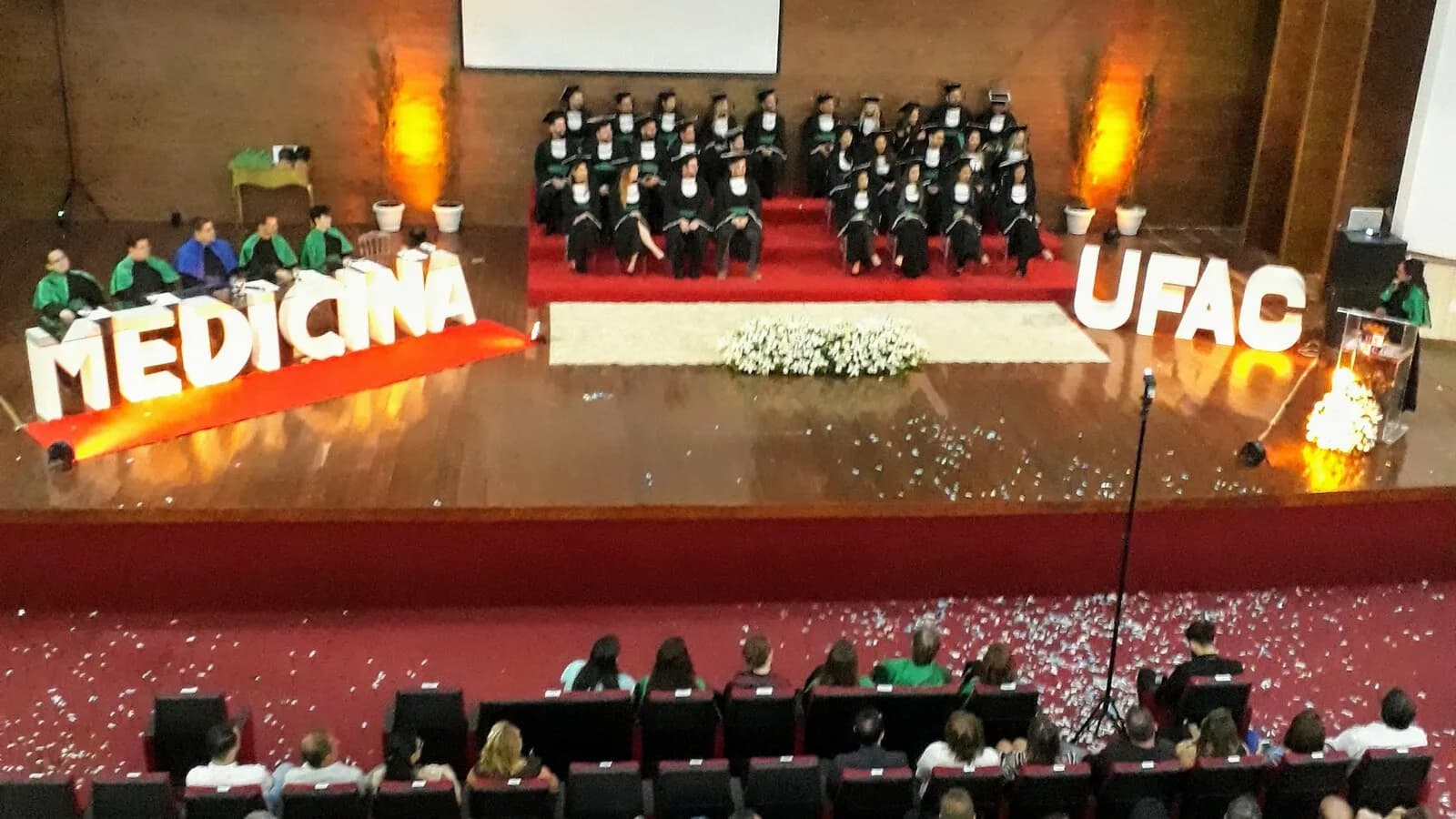 Formandos de beca no palco durante cerimônia de colação de grau em auditório, com plateia ao fundo