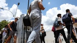 Candidatos com mochilas entram em local de prova em fila, sob céu com nuvens