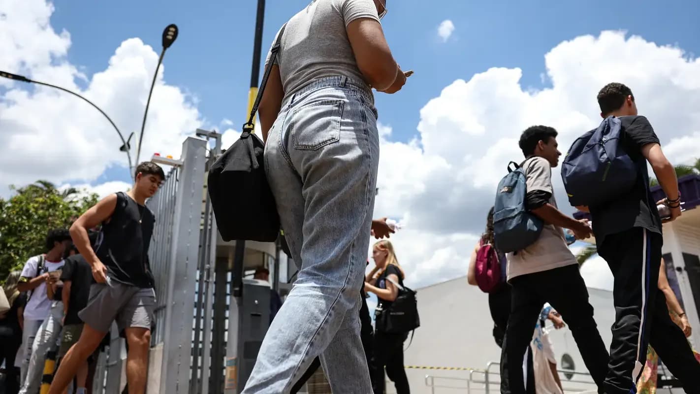 Candidatos com mochilas entram em local de prova em fila, sob céu com nuvens