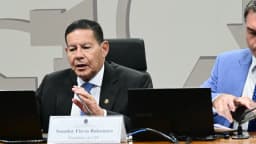 Senador Flávio Bolsonaro discursa sentado à mesa da Comissão de Segurança Pública do Senado, com placa de identificação como presidente da comissão