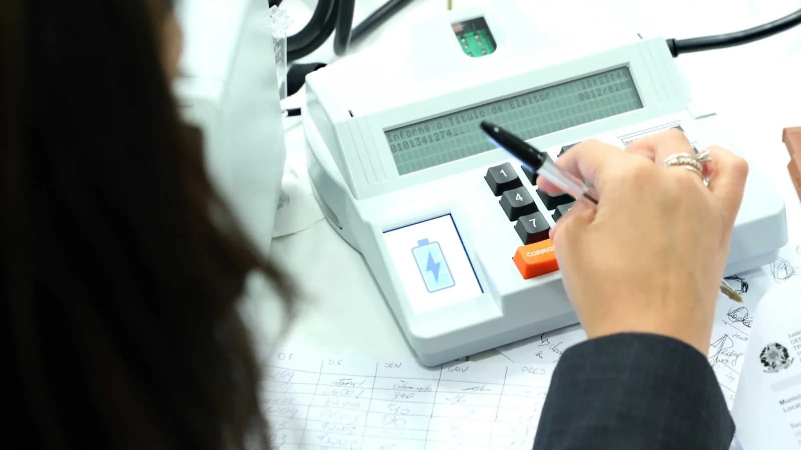 Mesária utiliza urna eletrônica durante teste de segurança, com formulários de apuração ao lado