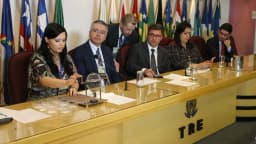 Mesa de sorteio de urnas eletrônicas em tribunal eleitoral, com integrantes da comissão ao redor de uma urna sobre a mesa