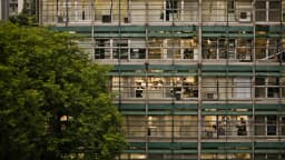 Fachada de prédio corporativo iluminado à noite, com funcionários visíveis em seus postos de trabalho