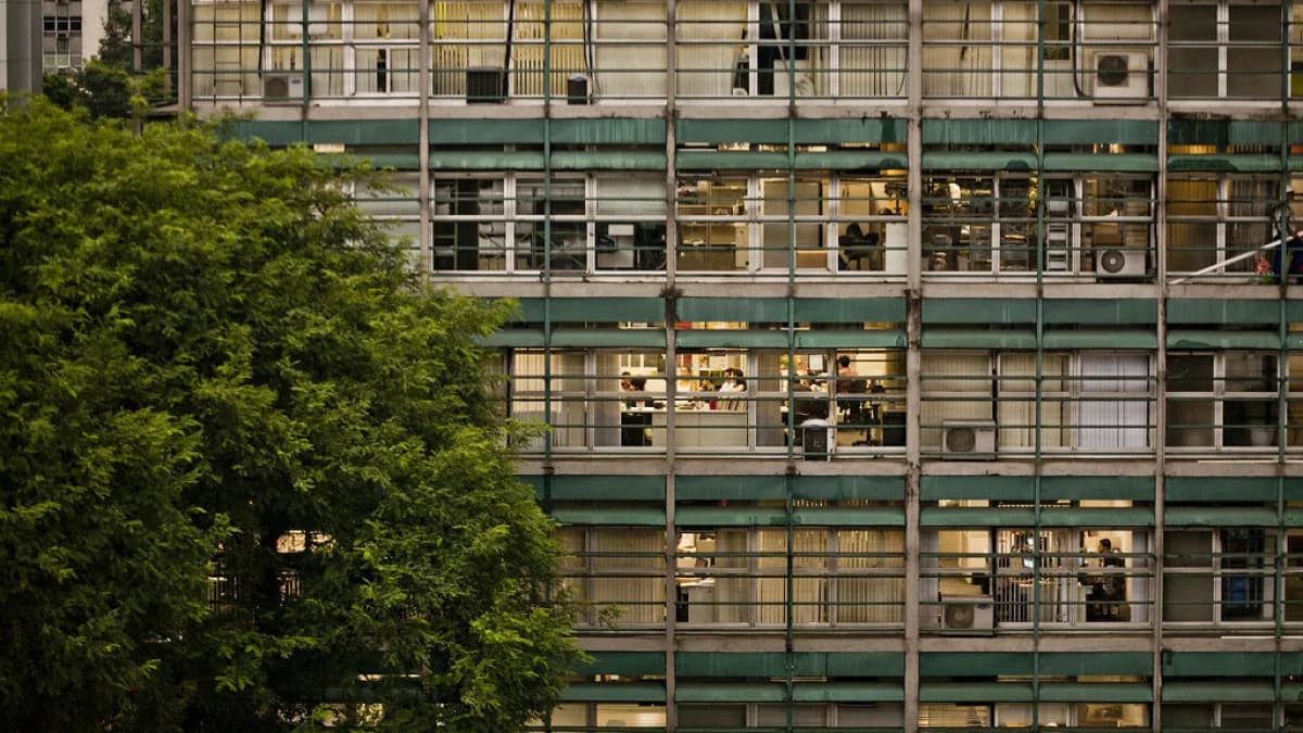 Fachada de prédio corporativo iluminado à noite, com funcionários visíveis em seus postos de trabalho