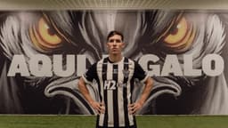 Thiago Borbas com a camisa listrada do Atlético-MG, mãos na cintura, diante de painel com o mascote do clube e os dizeres Aqui é Galo