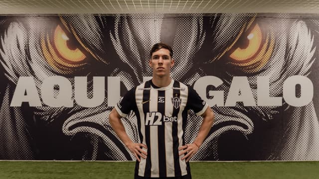 Thiago Borbas com a camisa listrada do Atlético-MG, mãos na cintura, diante de painel com o mascote do clube e os dizeres Aqui é Galo