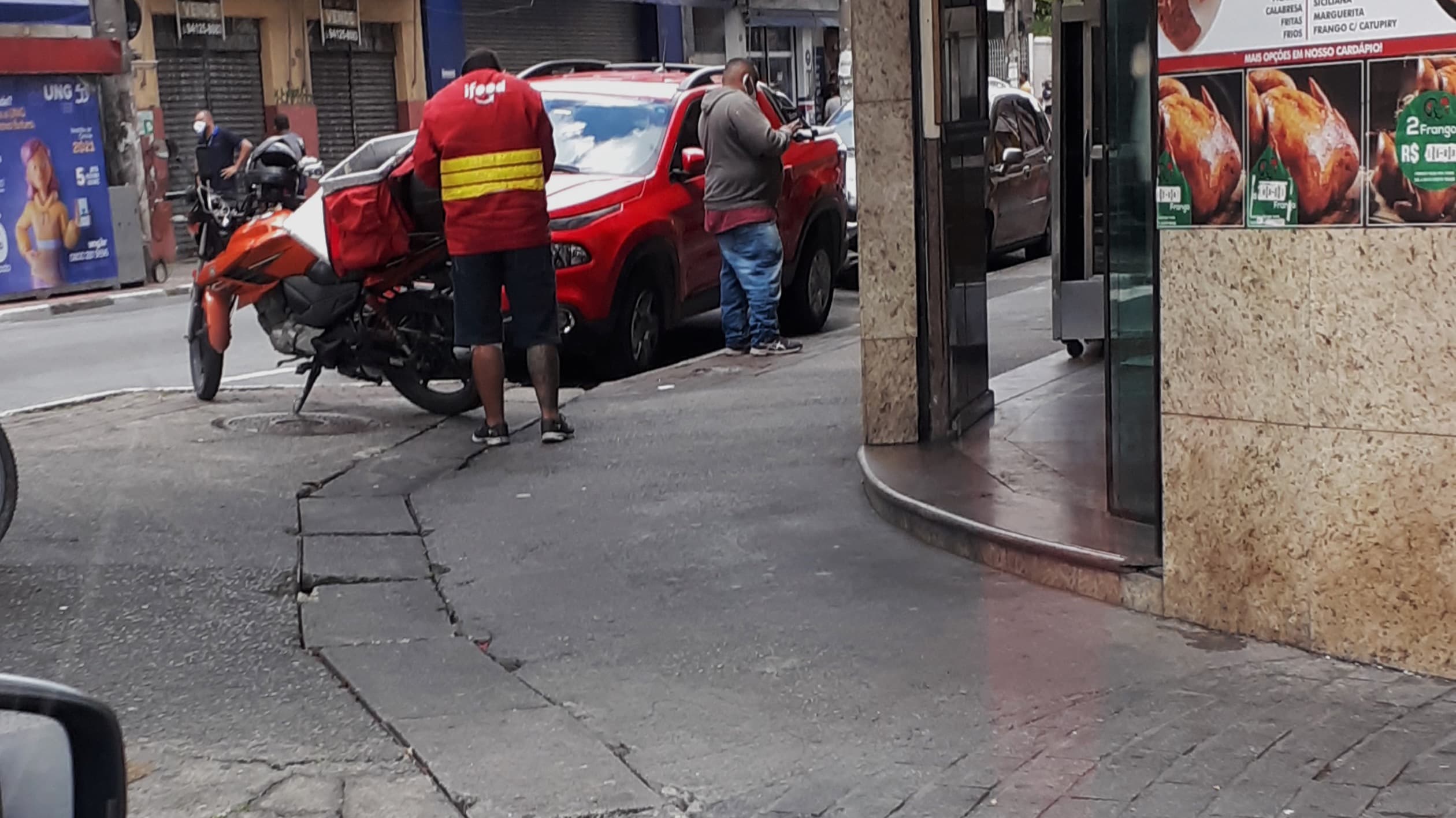 Entregador de aplicativo do iFood, de costas, ao lado da moto com a bag térmica vermelha, em rua de São Paulo