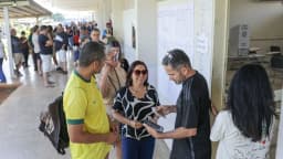 Fila de eleitores aguarda para votar em seção eleitoral, com cabine de votação ao fundo