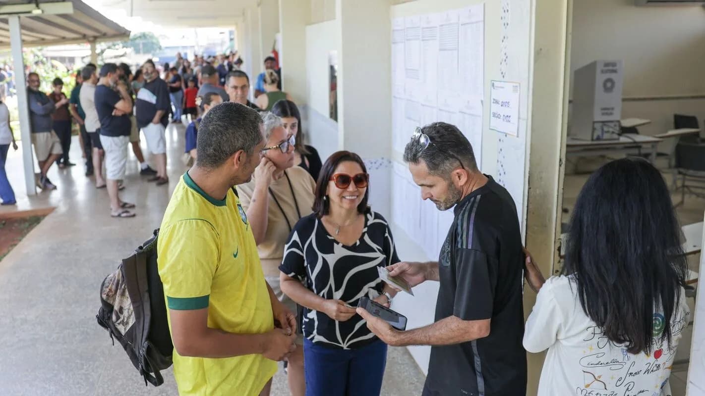 Fila de eleitores aguarda para votar em seção eleitoral, com cabine de votação ao fundo