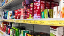 Foto de contexto de prateleiras de farmácia com caixas de medicamentos isentos de prescrição e etiquetas de preço, registrada em Brasília