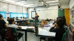 Professora em pé à frente do quadro branco de uma sala de aula de escola pública, com alunos sentados nas carteiras, em foto de arquivo