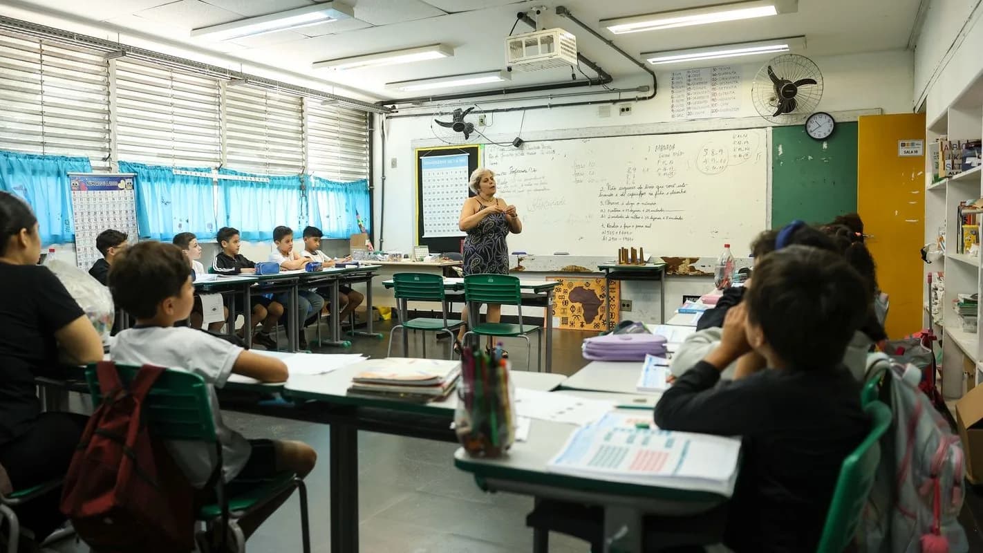 Professora em pé à frente do quadro branco de uma sala de aula de escola pública, com alunos sentados nas carteiras, em foto de arquivo