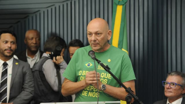 Luciano Hang discursa ao microfone, vestindo camiseta verde da Havan, em evento com bandeira do Brasil ao fundo