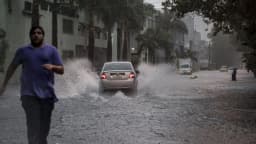 Homem corre sob chuva forte em rua alagada, com carro passando ao fundo