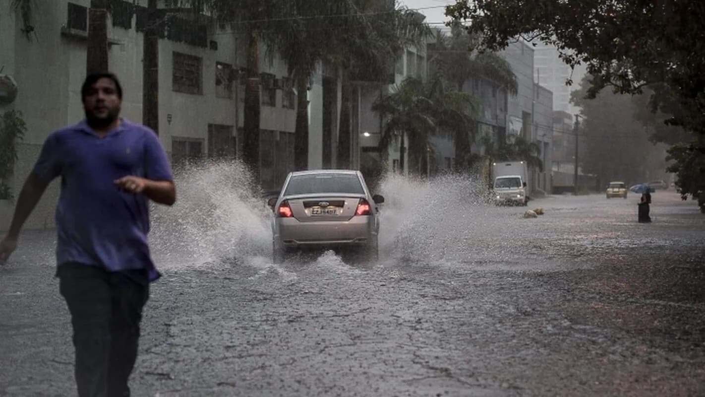 Homem corre sob chuva forte em rua alagada, com carro passando ao fundo