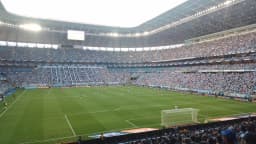Torcida e jogadores durante partida na Arena do Grêmio, estádio lotado visto da arquibancada