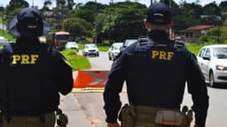 Dois policiais rodoviários federais de costas em fiscalização numa rodovia, com veículos ao fundo