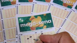Volantes da Mega-Sena espalhados sobre uma mesa, com uma mão marcando dezenas com caneta; imagem de arquivo