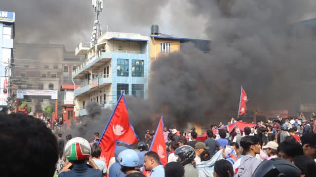 Manifestantes da Geração Z em protesto no Nepal, com bandeiras e fumaça ao fundo