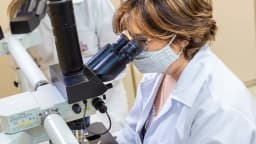 Pesquisadora de jaleco branco observa amostra em microscópio de laboratório
