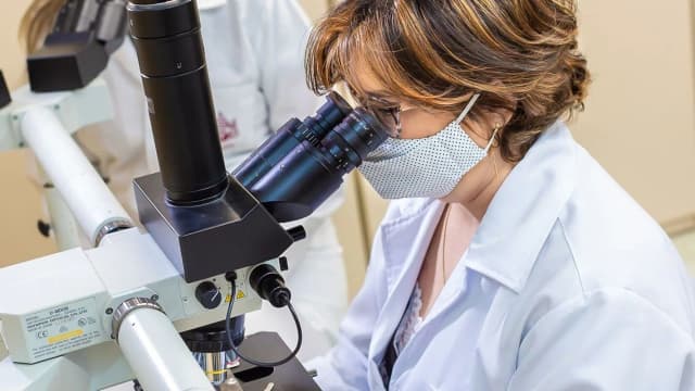 Pesquisadora de jaleco branco observa amostra em microscópio de laboratório