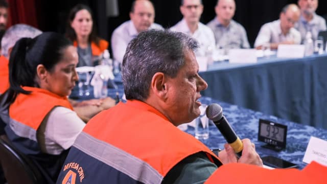 Governador Tarcísio de Freitas fala ao microfone em reunião de secretariado, vestindo colete da Defesa Civil