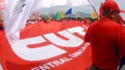 Sindicalistas seguram um bandeirão vermelho da CUT durante manifestação em Brasília; à direita, um homem de camisa e boné vermelhos aparece de costas, em imagem de arquivo