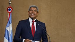 Jerônimo Rodrigues, de terno azul e gravata vermelha, sorri ao microfone ao lado da bandeira da Bahia, em foto de arquivo